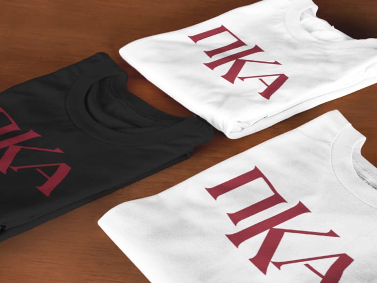 Customizable fraternity t-shirts with Greek letters ΠΚΑ in red, displayed in black, white, and gray color options on a wooden surface.