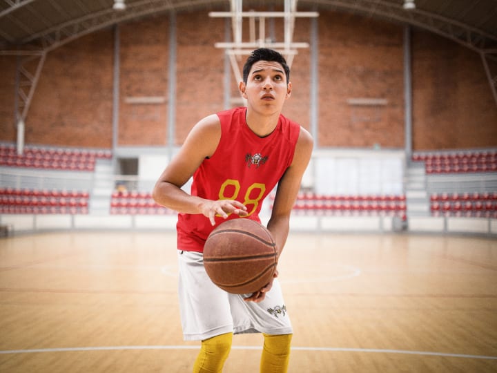  basketball player wearing a red jersey holds a basketball on an indoor court with high ceilings and empty bleachers.