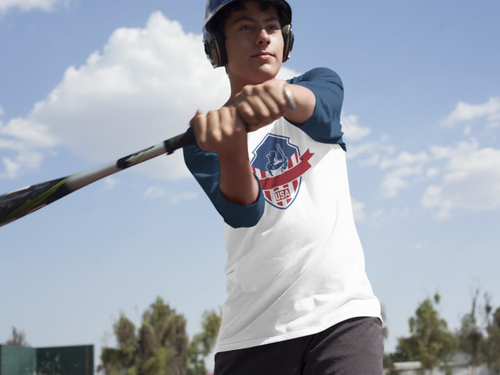 A baseball player in a long-sleeve shirt prepares to swing a bat outdoors, with a bright blue sky in the background.
