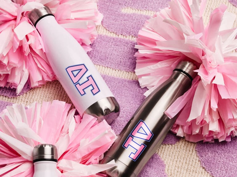 Customizable stainless steel water bottles with Greek letters ΑΓ, surrounded by pink pom-poms, placed on a purple patterned rug, highlighting a range of Greek life merchandise.