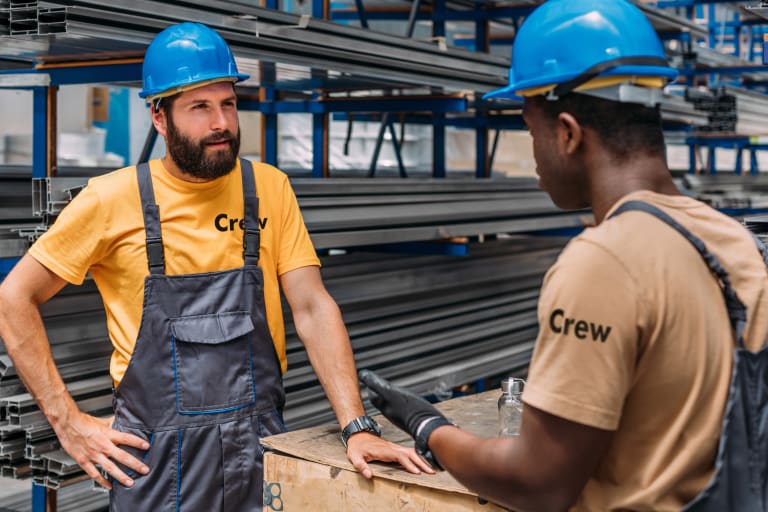Crew" T-shirts and blue hard hats have a discussion at a worksite. One worker, with a beard and overalls, leans on a wooden box while listening to the other, who is wearing gloves and gesturing with his hands. Metal beams are stacked on shelves in the background.