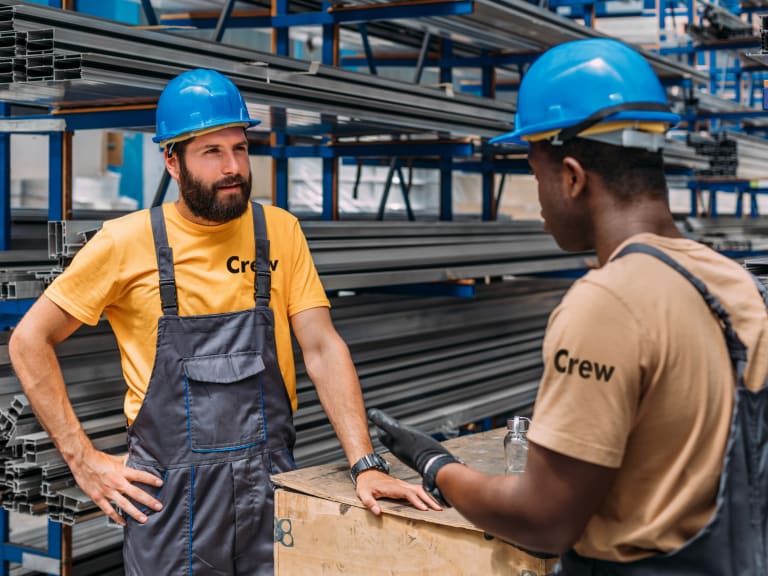 Crew" T-shirts and blue hard hats have a discussion at a worksite. One worker, with a beard and overalls, leans on a wooden box while listening to the other, who is wearing gloves and gesturing with his hands. Metal beams are stacked on shelves in the background.