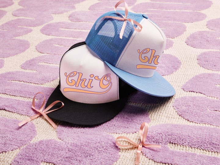 Variety of sorority trucker caps with Greek letters, arranged on a pink patterned background.