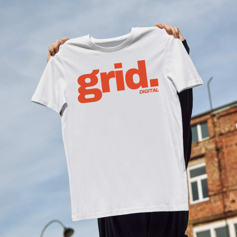 A person holds up a custom branded white T-shirt featuring bold red text that reads "grid. DIGITAL" against a backdrop of a brick building and a clear blue sky.