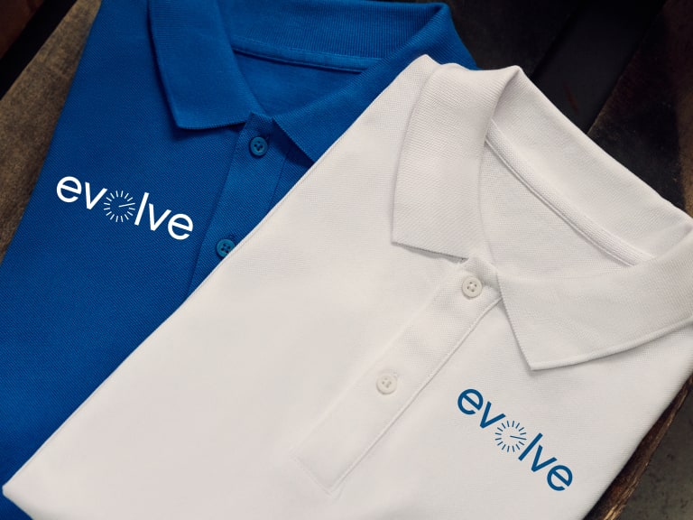 Two embroidered polo shirts, one blue and one white, are laid out side by side. Both feature the "evolve" logo in white and blue text with a circular design element.