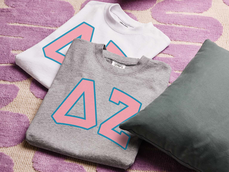 Customizable sorority t-shirts featuring Greek letters ΔΖ in pink and blue, neatly folded on a purple patterned rug, showcasing different color options.