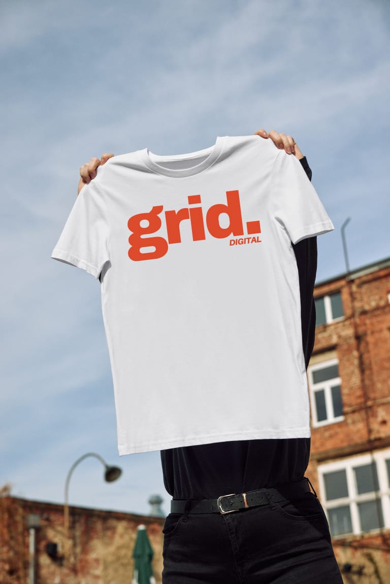 A person holds up a custom branded white T-shirt featuring bold red text that reads "grid. DIGITAL" against a backdrop of a brick building and a clear blue sky.